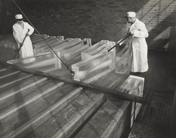 Two men dressed in lab coats and hats are holding pick axes to move ice bricks out from metal containers in the foreground. A line of ice bricks are behind them.