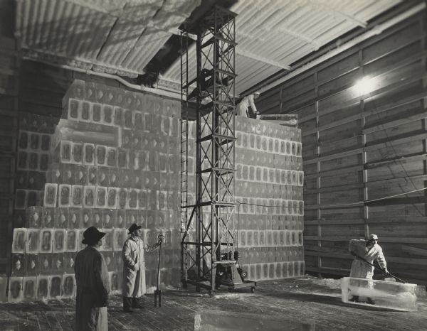 Four men are in a large room with a high ceiling. Three of the man are on the floor working to move ice bricks towards the stack of ice in the background. An elevator to move the ice is in the center, and one man is standing on top of the wall of ice up near the ceiling moving an ice brick into place.