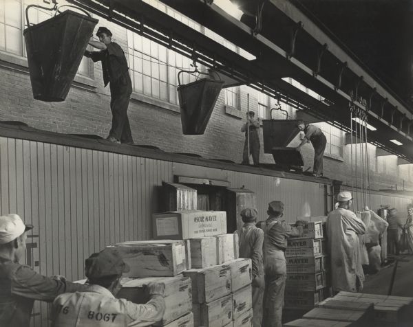 Men wearing overalls, jackets, hats, and gloves are pushing carts of wooden and cardboard crates which read: "Oscar Mayer, Approved, Fine Foods since 1883," toward the opening of a shipping (train) car. Standing and working on the roof of the shipping car are men moving large hanging chutes, opening doors in the roof, and using pick axes to load the cars with ice.