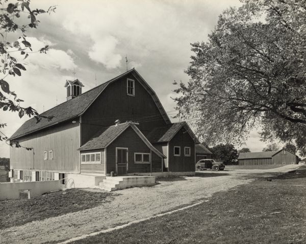 View across driveway towards barns and other outbuildings at the Oscar Mayer farm. There is an automobile parked near the barn.
