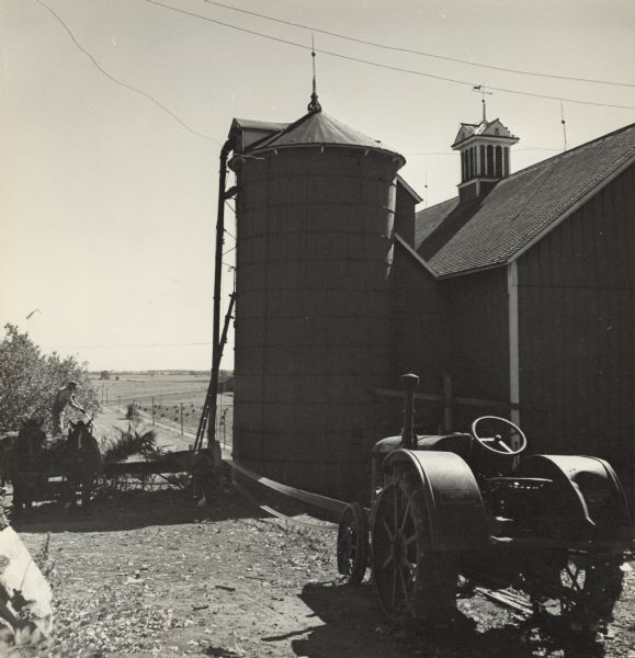 In the foreground on the right is a tractor belt-driving what may be an ensilage cutter. A man is standing on a horse-drawn wagon moving silage into the cutter to fill the silo next to a barn. There is a lightning rod on top of the silo, and a weather vane on top of a roof vent on the barn. In the background is a field dotted with corn shocks. 