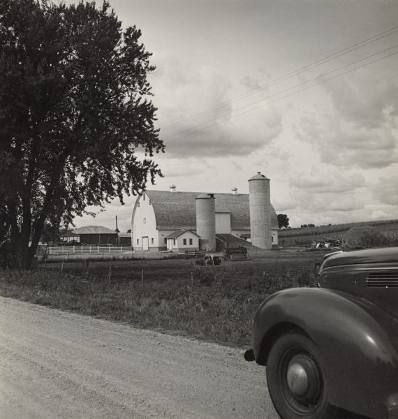 View from road, with the front of an automobile on the right, towards a farm. Cows and pigs are in a pasture, and two silos are next to the barn. 