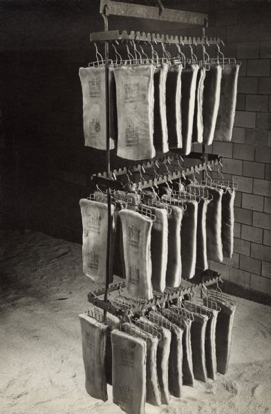 A rack is hanging from the ceiling above a floor spread with salt. Slabs of meat are hanging from hooks in three tiers on the rack to cure. The slabs are stamped with "Oscar Mayer Approved."