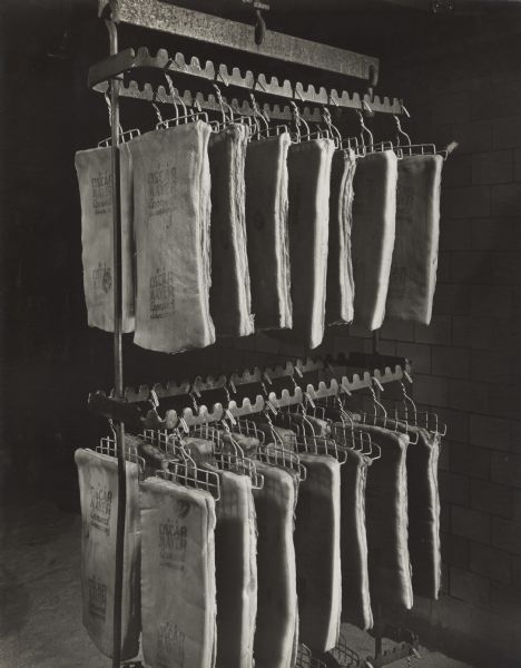 A rack is hanging from the ceiling above a floor spread with salt. Slabs of meat are hanging from hooks in three tiers on the rack to cure. The slabs are stamped with "Oscar Mayer Approved."