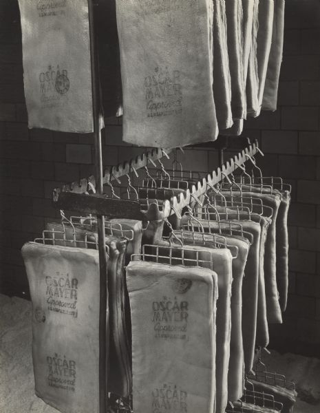 Close-up of meat hanging to cure on a metal rack hanging from the ceiling. The slabs are stamped with "Oscar Mayer Approved."