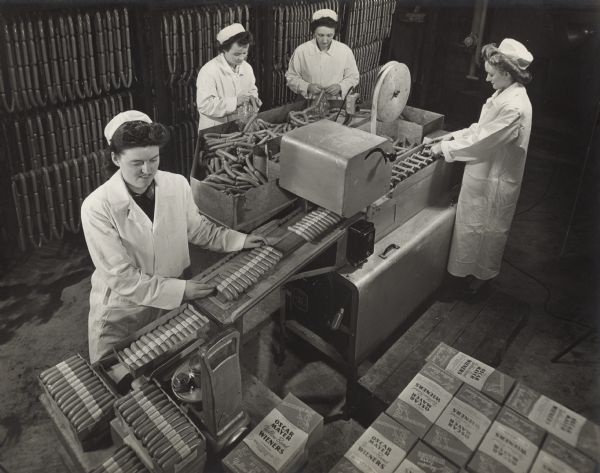 Slightly elevated view of four women standing near a conveyor belt and racks of wieners. They are wearing long coats and hats, and are labeling and packing Yellow Band Wieners. Two women in the background are separating lengths of weiners from a sheathed chain into individual wieners. The woman on the right is placing the individual wieners onto the conveyor belt to move them to a machine that is labeling the wieners in groups of 13. The woman on the left is placing the packaged wieners into a box. Boxes of packaged wieners are stacked in the right foreground. 