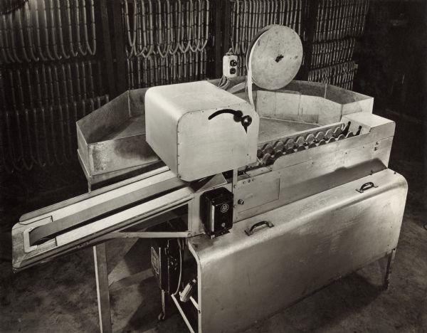 View of an Oscar Mayer wiener indexing and labeling machine, which has an attached sorting and holding section. Links of wieners are hanging on racks in the background.