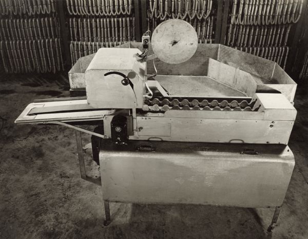 Side view of an Oscar Mayer wiener indexing and labeling machine, with an attached sorting and holding section. Links of wieners are hanging on racks in the background.