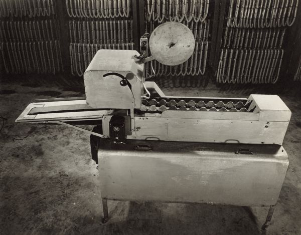 Side view of an Oscar Mayer Wiener wiener indexing and labeling machine, without the attached sorting and holding section. Links of wieners are hanging on racks in the background.