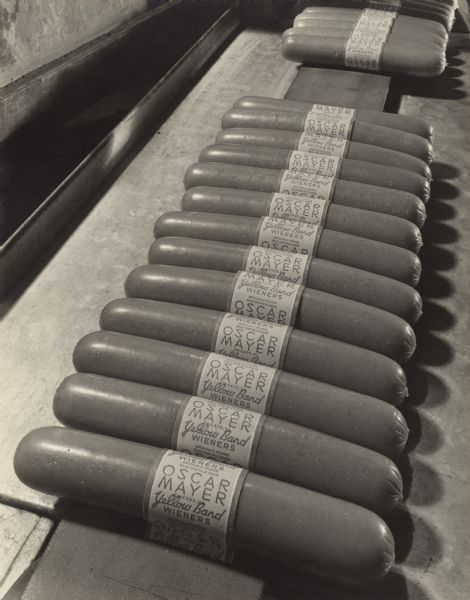 Close-up view of labeled Yellow Band Wieners in grouped in row of thirteen after exiting the labeling machine. The label reads: "Oscar Mayer, Chicago, Ill., Yellow Band Wieners, Artificially colored, Dried skim milk added."