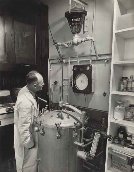 A male Oscar Mayer employee wearing a long white lab coat is standing next to a pressure cooker looking at a circular record keeping device attached to the wall.