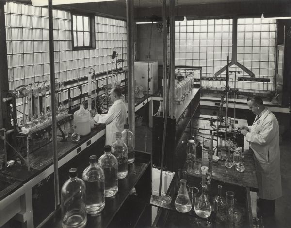 Elevated view of two men in a chemistry lab with glass block walls. The men are standing in front of two different wet bench lab tables, and are working on food chemistry tests and experiments for the Oscar Mayer Co. The room is filled with shelves holding glass bottles, beakers, stands, and gas burners. 