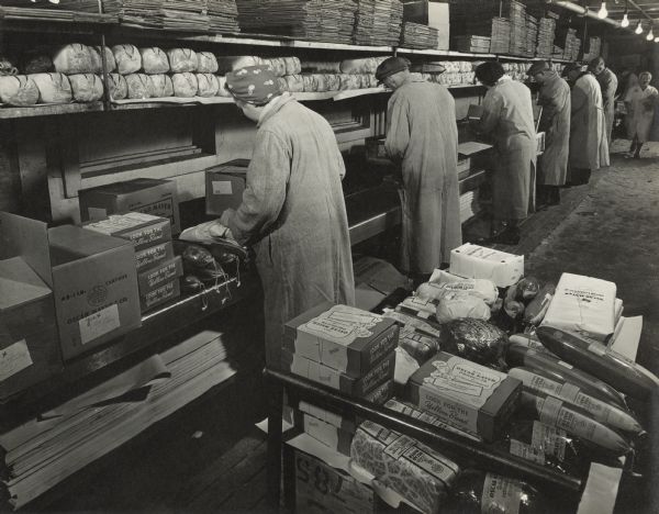Employees are standing in a row at a long counter filling different types of packaged meat into shipping boxes. In the foreground on a cart are boxes and packages of meat.