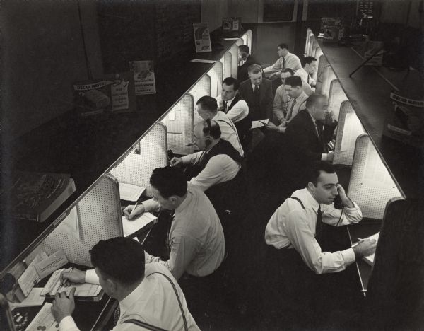 Elevated view of men sitting at a row of cubicles. Some of the men are talking telephones, and other men are writing on legal pads. Oscar Mayer advertisements are posted above the desks, and one reads: "Oscar Mayer Ham, Yellow Band, Mild, Honey Cured and selected for tenderness." 