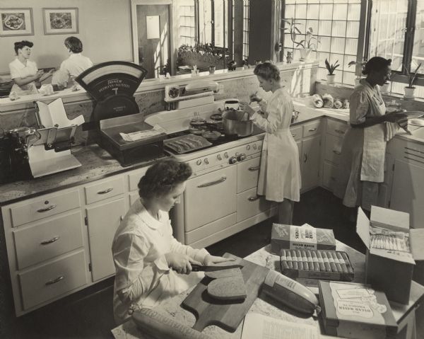 Slightly elevated view of five women researchers performing various aspects of food research and related activities in a test kitchen and laboratory at the Oscar Mayer Company. A woman is sitting at a table cutting meat in the foreground. Behind a low wall in the background two women are working at desks.