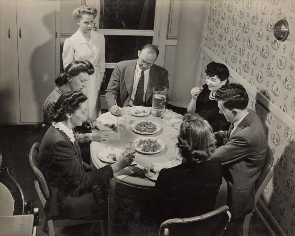 Slightly elevated view of men and women sitting around a table, sampling slices of Oscar Mayer meat products in the Oscar Mayer laboratory. A women in a long white coat is standing near the table watching the group of people. Each person sitting at the table is writing down an assessment of each sample of hotdog.
 
