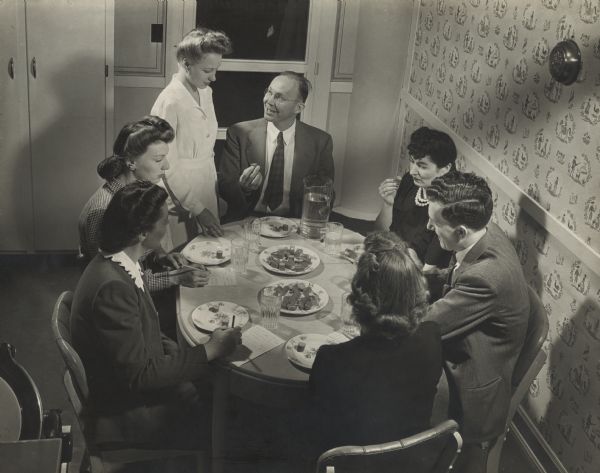 Slightly elevated view of men and women sitting around a table, sampling slices of Oscar Mayer meat products in the Oscar Mayer laboratory. A women in a long white coat is seen standing near by looking down and talking to man sitting at the table presumably introducing the dish. Individuals around the table write down critique of the sample of hotdog with a pencil and paper. 