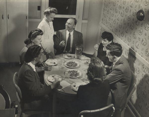 Slightly elevated view of men and women sitting around a table, sampling slices of Oscar Mayer meat products in the Oscar Mayer laboratory. A women in a long white coat is seen standing near by looking down and talking to man sitting at the table presumably introducing the dish. Individuals around the table write down critique of the sample of hotdog with a pencil and paper.
 