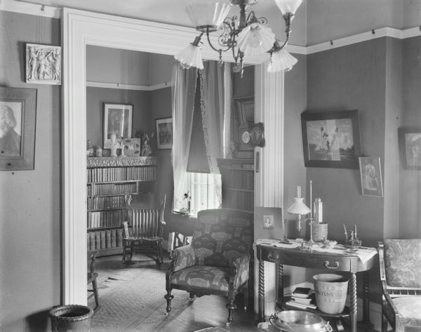 Interior of Carl A. Johnson residence, showing an adjoining library and sitting room with upholstered arm chairs and wooden side chairs, table, lace drapery, handwoven carpets, lamps and chandeliers, paintings, sculpture, and Native American baskets. The home was located on 142 East Gilman Street.