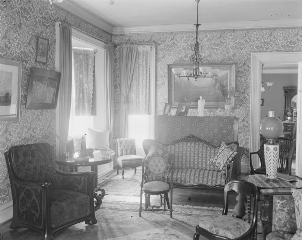 Interior of Carl A. Johnson residence, showing ornate parlor room with upholstered furniture (couch, arm chair and side chairs), end tables, patterned wallpaper, drapery, handwoven carpets, lamps and chandeliers, paintings, and sculpture. The home was located on 142 East Gilman Street.