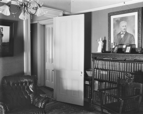 Interior of Carl A. Johnson residence, showing ornate study room with upholstered furniture (arm chair, side chair, end table and bookcase), handwoven carpet, chandeliers, painting, sculpture, Native American basket and framed mirror. The home was located at 142 East Gilman Street.