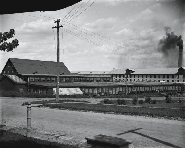Street view showing the back side of Gisholt machine works buildings located in the 1400 block at the corner of E. Mifflin and N. Dickinson streets. The intersection is of East Dayton and North Dickinson streets. The brick plant in the background, spewing smoke from a coal chimney, is Fuller & Johnson.