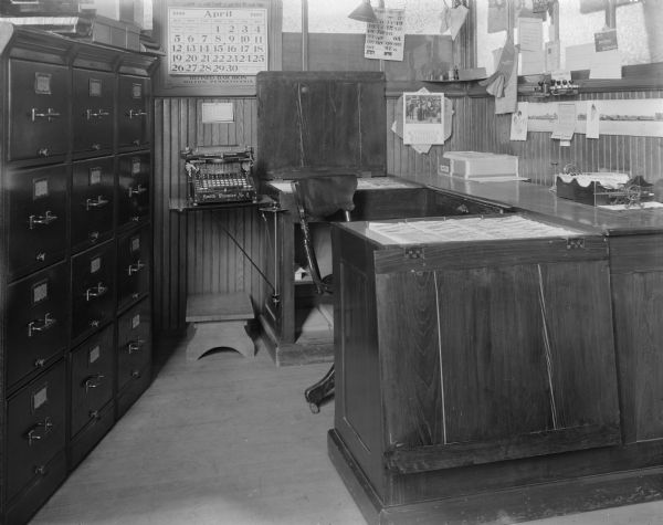 Interior of office with wooden rolling chair, filing cabinets, calendars (for April 1908), Smith Premier No. 2 typewriter, and rubber hand stamps. On the wall is a city panorama taken from offshore, including a steamboat.