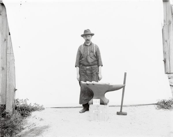 Straight-on standing portrait of a Gisholt laborer in work apron, suspenders, boots and brimmed hat. Taken outdoors before a white sweep, he is standing with his arms at his side behind an anvil placed on a wooden platform. An upside-down sledgehammer rests against the anvil. There are five metal bars (painted white to simulate hot metal) lined up on the anvil for forging.