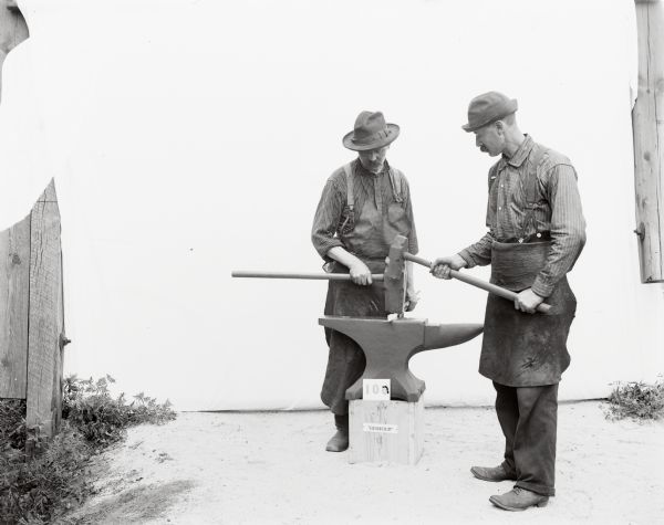 Portrait oftwo Gisholt laborers. One man is in profile and the other is facing front. They are wearing work aprons, suspenders, boots and brimmed hats. Taken outdoors before a white sweep, they are standing on opposite sides of the anvil placed on a wooden platform. The man on the right is holding a smaller hammer to demonstrate how workers forge hot metal into shape by pounding, while the man on the left is holding the metal bar in place with a sledgehammer in one hand and tongs in the other hand.