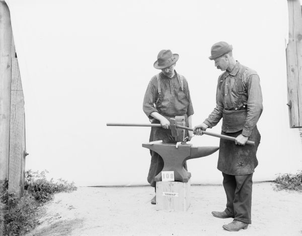 Portrait of two Gisholt laborers. One man is in profile and the other is facing front. They are wearing work aprons, suspenders, boots and brimmed hats. Taken outdoors before a white sweep, they are standing on opposite sides of the anvil placed on a wooden platform. The man on the right is holding a smaller hammer to demonstrate pounding a white-hot metal band into shape, while the man on the left is holding the band in place with a sledgehammer in one hand and tongs held in the other hand.