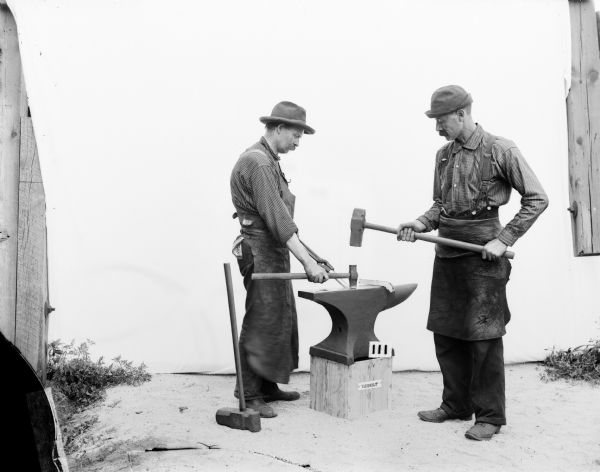 Portrait of two Gisholt laborers. One man is in profile and the other is in semi-profile. They are wearing work aprons, suspenders, boots and brimmed hats. Taken outdoors before a white sweep, they are standing on opposite sides of the anvil placed on a wooden platform. The man on the right is holding a hammer to demonstrate pounding a white-hot metal band into shape, while the man on the left is holding the band in place with tongs in one hand to alternately pound with a smaller hammer with the other hand.