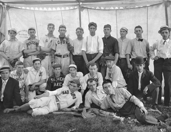 Group portrait of baseball players, wearing jerseys from Madison collegiate and amateur teams including the Badgers, Crescents and Silver Lake. The players are under a tent provided by the Madison Tent and Awning Company owned by W.G. Kropf of Madison, Wisconsin. One man is holding a bat, and the others are holding mitts. 