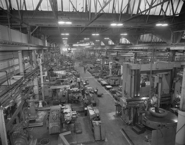 Elevated view of factory floor at the Gisholt plant, located in the 1200 and 1300 blocks of East Washington Avenue. There is a bridge crane, rows of work stations, and several operators at stations manning automated machinery.