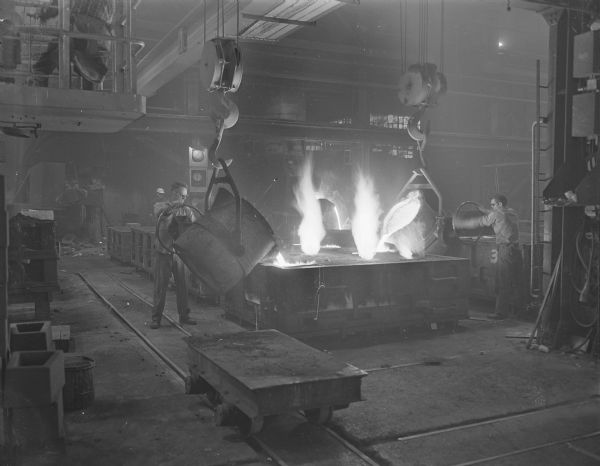 View of two workers pouring molten metal on the floor of the Gisholt plant. There are two massive chain hoists, suspended from a bridge crane, that are holding the vats of liquified metal being poured into molds. Tracks in the floor are for moving supplies and casts. 