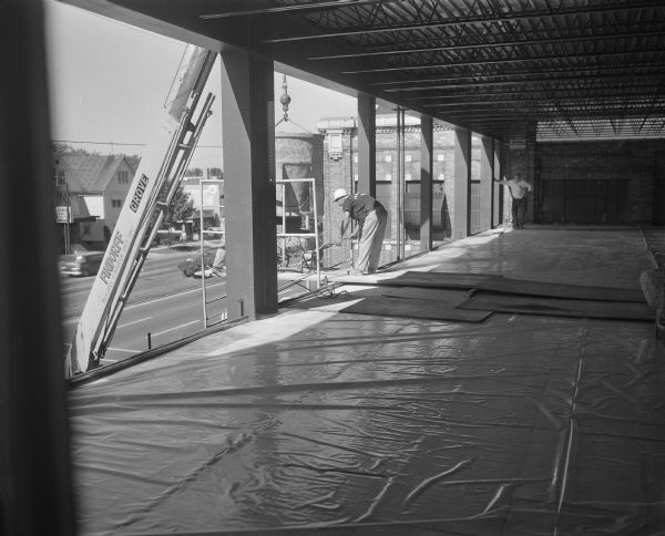 View of construction of the skywalk across Baldwin Street at the Gisholt plant, facing East Washington Avenue. Two workers, one obscured by a beam, are unloading a wheelbarrow and other materials from a scaffolding lift, and a Findorff crane is hoisting a bucket above the scaffolding. The offices of Messner Incorporated and a realtor are located across the street.
