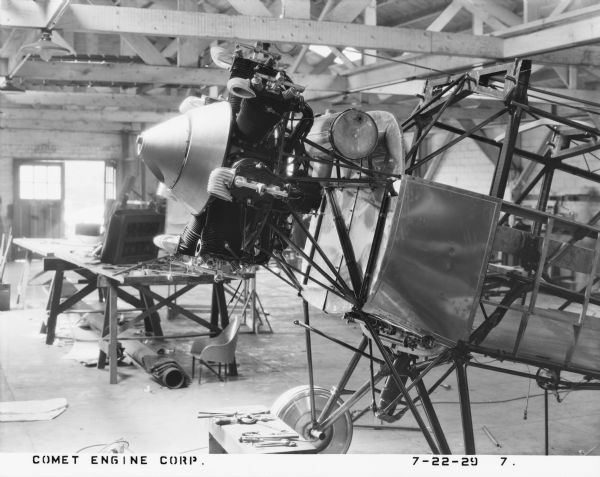 Left side view of the 7-cylinder Comet engine fitted to the nose of an aircraft under construction. The fuselage is standing on the factory floor. A work table and exit doors to the outside are in the background. 
