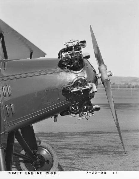 Profile view of a 7-cylinder Comet air-cooled radial piston engine, designed by the Comet Engine Corporation on an unidentified make of aircraft at the Oakland landing strip. The photograph pictures the front end of an airplane showing a partial wing, wheel and strut, engine on nose and wooden propeller.