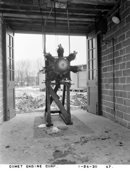 View from front of a 7-cylinder Comet airplane engine undergoing assembly. The engine is mounted to a wooden stand, located in a brick manufacturing building, and shows its crankcase and component parts like nose cowling, valves cylinders, and spark plugs. The doors in the background are open to a snowy landscape.