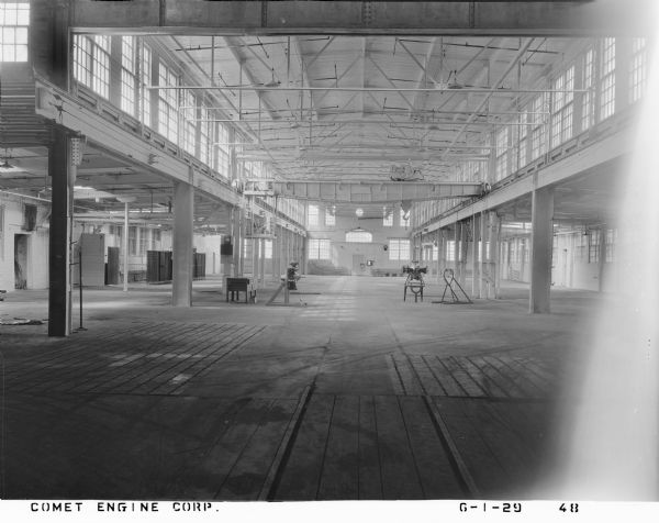 Two 7-cylinder Comet engines assembled with cams are set up on a factory floor. Supported in opposing engine stands vertically and horizontally, they are under an overhead crane that reads: "Whiting Foundry Equipment Co. of Chicago" on the manufacturer's plate. The interior of the factory floor is sparse with production, and shows architectural details like steel girders, electrical lighting and wires, doors, windows on first and second stories, ceiling sprinklers, and floor tracks. 