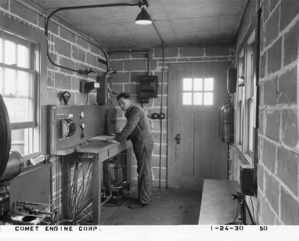 View towards a factory man working at a plant that assembled Comet engines for aircraft. The man is standing at a desk fitted with instruments in a narrow brick office space. The opposite wall has a Foamite fire extinguisher mounted to the wall and a candlestick telephone on a desk.