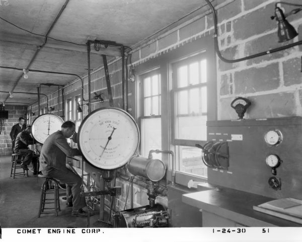 View of two factory men at work desks at a plant that assembled Comet aircraft engines. They are sitting on wooden stools between Korn scales in a narrow brick office space. A man wearing a suit and tie is standing over one worker in the background.