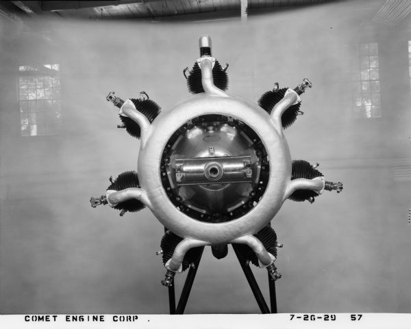 Straight-on view of the front crankcase of a 7-D Comet aircraft engine on a stand. It was made in front of a sheer backdrop inside a factory, with windows in the background. The engine had valves fore and aft, the latter bearing an exhaust valve for better cooling by air. The spark plugs are placed on the sides of the cylinder heads, made of a steel barrel. A design of the Comet Engine Corporation in 1928, the 7-D engine was manufactured on a Gisholt turret lathe in Madison, Wisconsin.