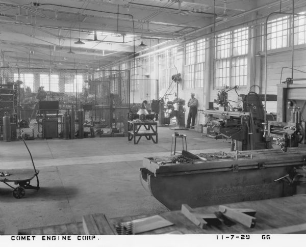 Interior view of a factory floor. Machinery by Whitcomb Manufacturing Company is in the right foreground, and a Comet 7-cylinder aircraft engine is in the center of the floor. A man, leaning against the wall by a machine, is looking towards the photographer. The room also includes an enclosed work space with tools, and is surrounded by a chain link fence. 