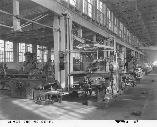 Interior of a factory floor, with several large machines for manufacturing aircraft engines, including Landis, Whitcomb Manufacturing Company and Gisholt. Men are working in the background (blurred from movement).