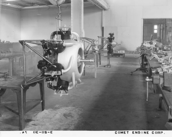 View inside a 7-D Comet aircraft engine manufacturing and assembly plant. An engine with nose cowling, propeller shaft and cylinders is supported by an engine stand and crane hook in the foreground. Another engine is on a stand in the background. Along the right are tables with tools and vices.  