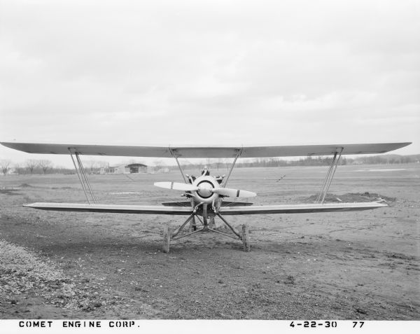 Front view of a Alexander Eaglerock A-12 aircraft fitted with the 7-RA Comet air-cooled radial piston engine with seven cylinders. The fixed-wing plane sitting on a dirt runway at an airport, with a hangar and homes in the background.
