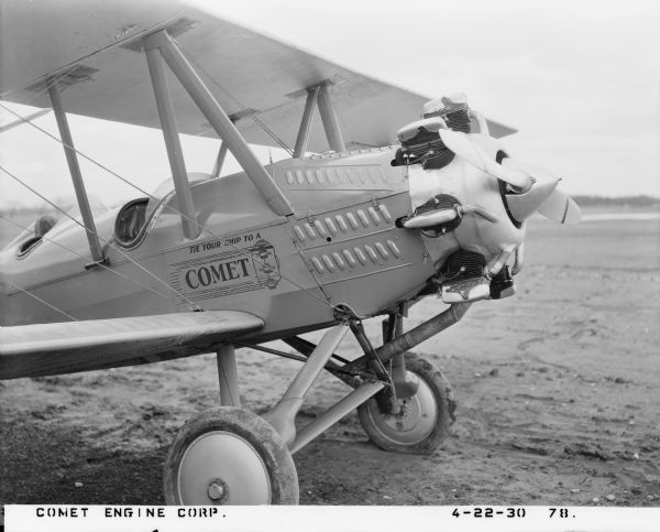 Three-quarter view from front right of an Alexander Eaglerock A-12 fixed-wing aircraft fitted with a 7-RA Comet engine. The plane is parked on an airstrip presumably at a hanger in Madison and was used for corporate advertising. Decal text on the plane body by the cockpit reads: "Tie your ship to a Comet."