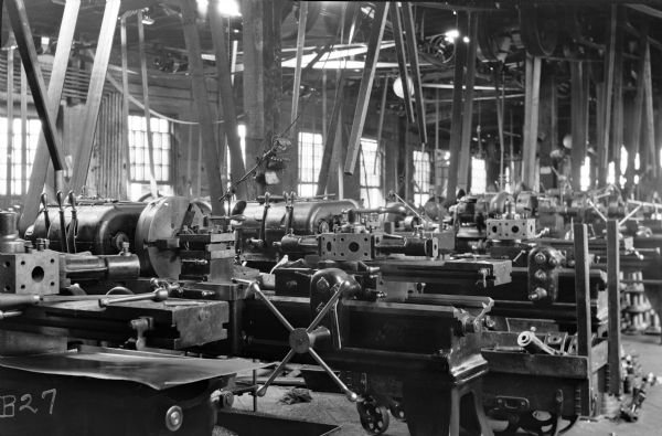 View of new turret lathe (center) manufactured by American Tool Works. It is standing on a factory floor flanked by other similar machines that are attached to rotary belts, suspended from the ceiling. Electrical light bulbs are strung above each work station. The factory room is lit by both natural light from the windows and electricity. 

The original envelope housing this photograph was inscribed with the following information: "They [the lathes] have a stop for cross carriage, same as Gisholt which you cannot see in view."