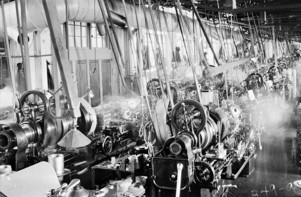 View of a battery of turret lathes in the "Gisholt Department" in a factory of the Peerless Auto Motor Car Company taken while machinery is in operation. The lathes flank each other that are attached to rotary belts, suspended from the ceiling. Several plant workers standing at their lathes in the aisles are blurred from motion. Hats and coats are on hooks on a wall lined with windows. 