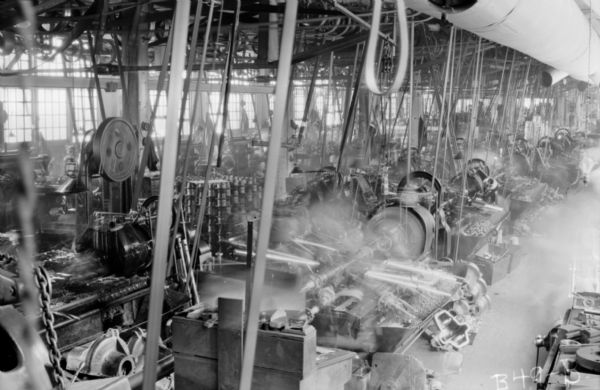 Elevated view of a room full of turret lathes in the "Gisholt Department" in a factory of the Peerless Auto Motor Car Company taken while the machinery is in operation. Lathes flank each other that are attached to rotary belts, suspended from the ceiling. Several plant workers standing at their lathes in the aisles are blurred from motion. Hats and coats are on hooks on a wall lined with windows.